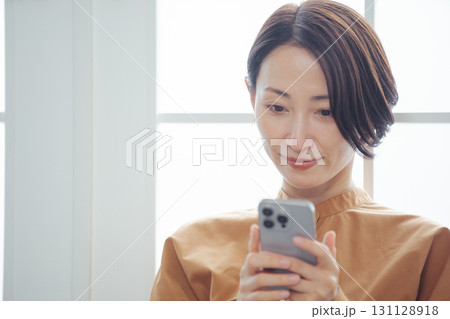 窓際でスマホを見ながら嬉しそうに微笑むミドル女性 窓際でスマホを見ながら嬉しそうに微笑むミドル女性 131128918