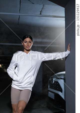 In a dark underground parking space a girl dressed in a white hoodie stands confidently. She leans against a pillar with a vehicle nearby creating a stylish urban scene. 131129417