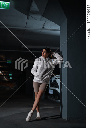 girl leans against a wall in an underground parking area. She wears a white hoodie and poses casually showcasing a relaxed yet confident vibe in low light. girl leans against a wall in an underground parking area. She wears a white hoodie and poses casually showcasing a relaxed yet confident vibe in low light. 131129418