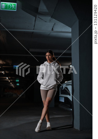 In the underground parking lot a girl wears a white hoodie and stands with confidence near a parked car. The dim lighting highlights her relaxed yet stylish look in an urban setting. In the underground parking lot a girl wears a white hoodie and stands with confidence near a parked car. The dim lighting highlights her relaxed yet stylish look in an urban setting. 131129420