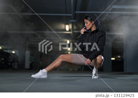 girl wearing a black hoodie crouches in a dimly lit underground parking lot. The atmosphere is enhanced by the surrounding fog and soft lights. She appears contemplative and stylish. girl wearing a black hoodie crouches in a dimly lit underground parking lot. The atmosphere is enhanced by the surrounding fog and soft lights. She appears contemplative and stylish. 131129425
