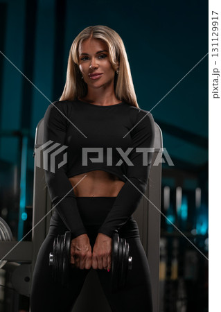 Blonde woman in a black workout outfit stands in a gym holding dumbbells and showcasing her strength. The scene captures her dedication to fitness and wellness during a training session. Blonde woman in a black workout outfit stands in a gym holding dumbbells and showcasing her strength. The scene captures her dedication to fitness and wellness during a training session. 131129917