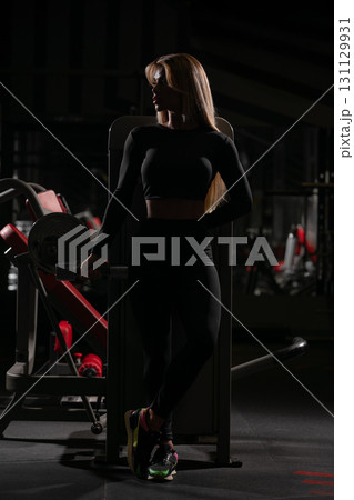 woman with blonde hair is standing in a dimly lit gym wearing a black workout outfit. She appears focused and relaxed as she prepares for her workout. woman with blonde hair is standing in a dimly lit gym wearing a black workout outfit. She appears focused and relaxed as she prepares for her workout. 131129931
