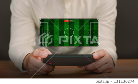Football fans watch a live football match on a smartphone screen with a green football field background. Concept of online sports streaming, digital entertainment, or live sports broadcast at home. 131130274