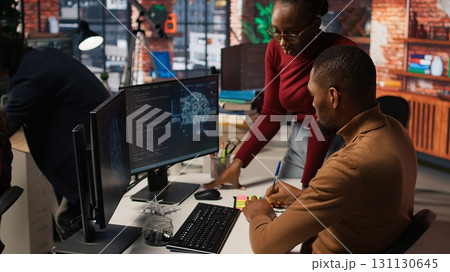 Teamworking startup engineers writing message on sticky notes while coding advanced AI algorithm. Coworkers in artificial intelligence Silicon Valley workspace leaving memo on PC screen, camera A 131130645