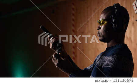 Portrait of happy shooting range customer reloading pistol, shooting targets, having fun. Close up of joyous man at shooting ground facility loading pistol, wearing protection goggles, camera A 131130678