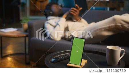 Black male remote worker laying on the couch next to chroma key display, enjoying leisure and free time for relaxation in his modern loft apartment with neon lights and a smart tv. Camera B. 131130730