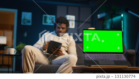 Young black adult uses his tablet next to green screen in his cozy modern apartment, laying on the couch and relaxing with his gadgets at home. Person with neon lights space, chill guy. Camera B. Young black adult uses his tablet next to green screen in his cozy modern apartment, laying on the couch and relaxing with his gadgets at home. Person with neon lights space, chill guy. Camera B. 131130774