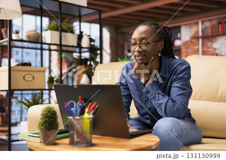 African american woman multitasking on a laptop in her stylish loft, fully connected to the internet while managing her freelance tasks and remote responsibilities. Browsing web at her home. African american woman multitasking on a laptop in her stylish loft, fully connected to the internet while managing her freelance tasks and remote responsibilities. Browsing web at her home. 131130999