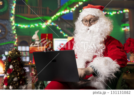 Person dressed as Santa Claus reading letters from kids on laptop in festive adorn workshop. Man portraying Christmas character answering emails received from children using notebook Person dressed as Santa Claus reading letters from kids on laptop in festive adorn workshop. Man portraying Christmas character answering emails received from children using notebook 131131082