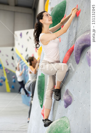 Girl is mastering climbing on training wall in gym. Woman holds on tightly to ledges and strives for top of bouldering route Girl is mastering climbing on training wall in gym. Woman holds on tightly to ledges and strives for top of bouldering route 131132845