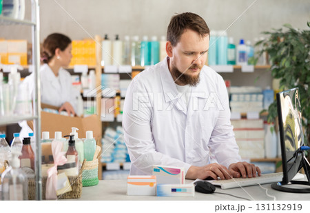 Man pharmacist uses computer at workplace. Girl assistant in background Man pharmacist uses computer at workplace. Girl assistant in background 131132919