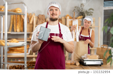 Guy seller in bakery hold out hand with phone, points at screen with finger 131132954