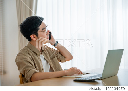 Happy young Asian man working on laptop in living room Happy young Asian man working on laptop in living room 131134070