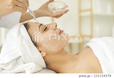 Female cosmetologist applying moisturizing mask with brush to a young woman in spa salon. 131134379