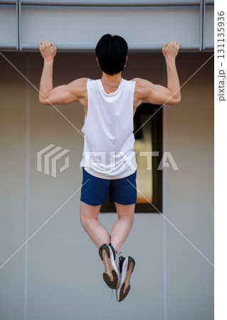 Full body of an asian man in white tank top doing a pull up on the horizontal bar in sports stadium. 131135936