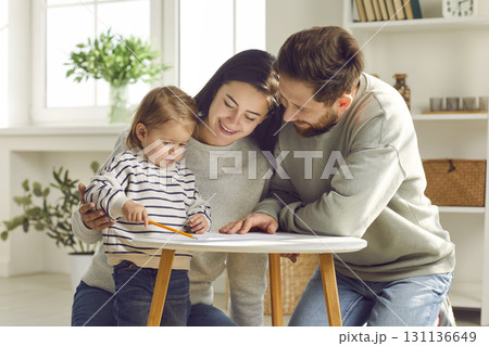 young married couple, together with their child daughter, sits on the floor and draws with a pencil on a white sheet of paper young married couple, together with their child daughter, sits on the floor and draws with a pencil on a white sheet of paper 131136649
