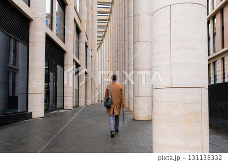 man in autumn coat walks under colonnade in business district of city, down town. man in full growth walks away from the camera. businessman, lawyer, manager, office worker man in autumn coat walks under colonnade in business district of city, down town. man in full growth walks away from the camera. businessman, lawyer, manager, office worker 131138332