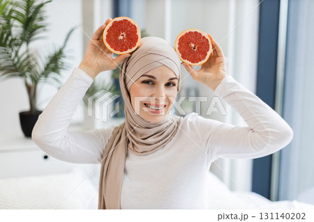 Cheerful young adult woman wearing hijab holding fresh grapefruit slices indoors, showcasing joy and creativity. Depicts a healthy lifestyle, humor, and aesthetic inspiration. 131140202