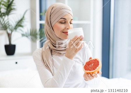 Smiling young Middle Eastern woman wearing hijab enjoys delicious yogurt and grapefruit. Demonstrates healthy lifestyle, natural skincare, and happiness. Home setting with natural light and plants Smiling young Middle Eastern woman wearing hijab enjoys delicious yogurt and grapefruit. Demonstrates healthy lifestyle, natural skincare, and happiness. Home setting with natural light and plants 131140210
