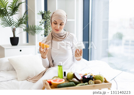 Young Muslim woman in her 20s sitting indoors preparing to use grapefruit and cream as part of her skincare routine, promoting natural wellness and self-care. 131140223