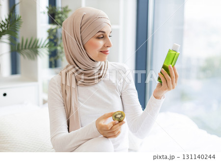 Young Muslim woman wearing hijab holding kiwi lotion bottle in indoor environment. Promotes natural skincare products and self-care practices focused on beauty, wellness, and hydration routines. 131140244