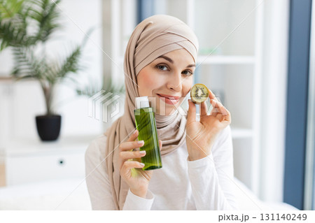 Young Muslim woman wearing headscarf holding kiwi fruit and skincare lotion bottle, smiling indoors with natural light, representing organic beauty products and skincare in minimalist environment. Young Muslim woman wearing headscarf holding kiwi fruit and skincare lotion bottle, smiling indoors with natural light, representing organic beauty products and skincare in minimalist environment. 131140249