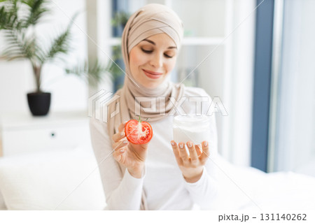 Young Muslim woman wearing beige hijab holding fresh tomato and skincare cream while smiling. Daylight ambience with indoors natural decor conveys skincare routine concept. Young Muslim woman wearing beige hijab holding fresh tomato and skincare cream while smiling. Daylight ambience with indoors natural decor conveys skincare routine concept. 131140262