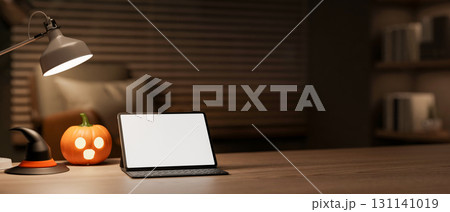 White screen tablet with halloween pumpkin lantern and witch hat under lamp on table in a dark room. 131141019