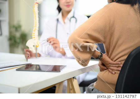 Patient holding lower back in pain while consulting doctor with spine model in clinic 131142797