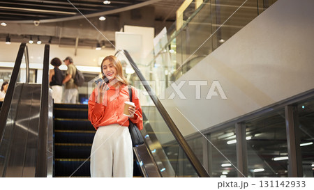 Young woman on escalator holding coffee cup and checking her messages in shopping mall Young woman on escalator holding coffee cup and checking her messages in shopping mall 131142933