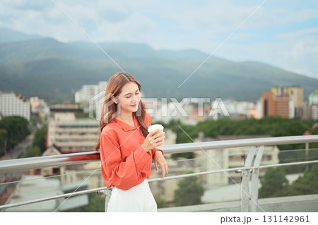 Relaxed businesswoman holding takeaway cup while standing on rooftop with city and mountain view Relaxed businesswoman holding takeaway cup while standing on rooftop with city and mountain view 131142961
