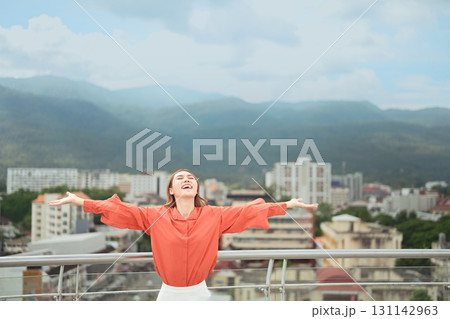 Cheerful woman standing on rooftop with arms wide open, smiling joyfully against cityscape and mountain view Cheerful woman standing on rooftop with arms wide open, smiling joyfully against cityscape and mountain view 131142963