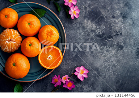 A vibrant arrangement of fresh oranges sits on a teal plate, accompanied by delicate pink flowers on a dark textured surface A vibrant arrangement of fresh oranges sits on a teal plate, accompanied by delicate pink flowers on a dark textured surface 131144456