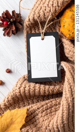 Fall Themed Product Tag on Beige Knitted Scarf with Autumnal Decorations on White Wooden Surface 131145586