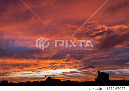 A vibrant sunset with shades of orange, pink, and purple clouds. Silhouettes of buildings are visible in the foreground against the colorful sky. 131147073