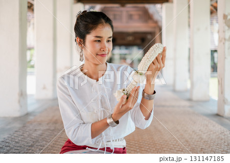 A woman in traditional Thai attire holds a floral garland, showcasing cultural heritage and intricate architecture. 131147185