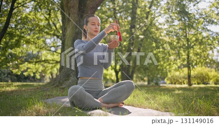 A young beautiful white girl doing yoga in the park and meditating in the lotus position among the trees in a clearing on a summer day and using a singing brass bowl A young beautiful white girl doing yoga in the park and meditating in the lotus position among the trees in a clearing on a summer day and using a singing brass bowl 131149016