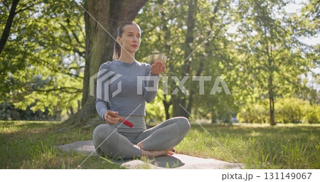 A young beautiful white girl doing yoga in the park and meditating in the lotus position among the trees in a clearing on a summer day and using a singing brass bowl A young beautiful white girl doing yoga in the park and meditating in the lotus position among the trees in a clearing on a summer day and using a singing brass bowl 131149067