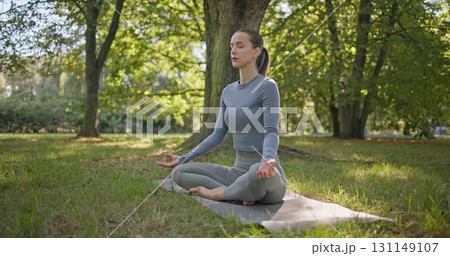 A young beautiful white girl practices yoga in the park and meditates in the lotus position among the trees in a clearing on a summer day 131149107