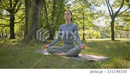 A young beautiful white girl practices yoga in the park and meditates in the lotus position among the trees in a clearing on a summer day 131149150