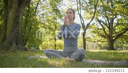 A young beautiful white girl practices yoga in the park and meditates in the lotus position among the trees in a clearing on a summer day 131149211