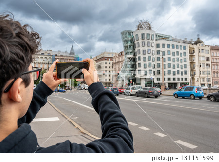 Prague, Czech Republic, August 7, 2023. A young Caucasian man photographs with his cell phone the fascinating three quarter view of the Dancing House: futuristic shapes and modern materials. Prague, Czech Republic, August 7, 2023. A young Caucasian man photographs with his cell phone the fascinating three quarter view of the Dancing House: futuristic shapes and modern materials. 131151599