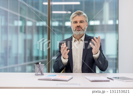 Senior businessman gesturing during an online video conference, discussing business strategies and communicating with clients or colleagues while working from a modern corporate office 131153021