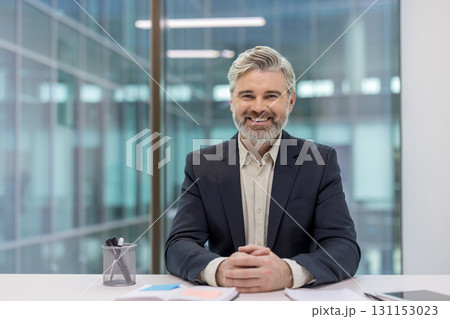 Mature bearded businessman sitting at his office desk, confidently smiling and looking at the camera, conveying professionalism, success, and trust in a modern corporate environment 131153023