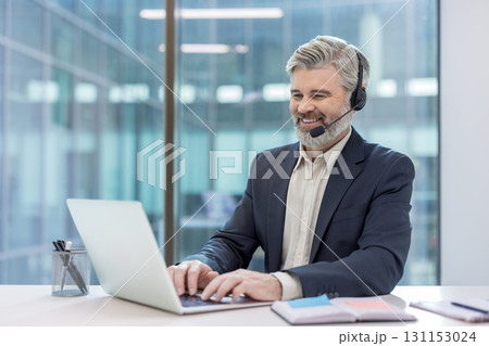 Mature businessman wearing a headset and smiling, actively typing on a laptop, providing customer service or technical support from a modern office environment Mature businessman wearing a headset and smiling, actively typing on a laptop, providing customer service or technical support from a modern office environment 131153024