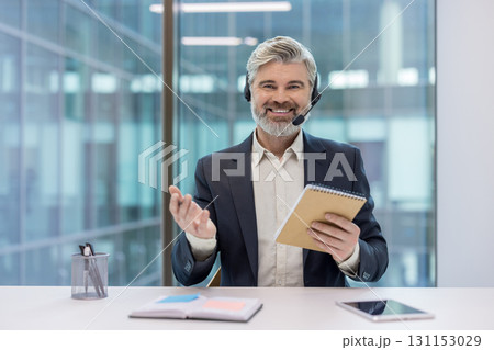 Mature call center agent in headset smiling at camera, holding notebook and gesturing while providing friendly professional customer support in a modern office setting 131153029