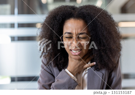 Close-up photo of an African American businesswoman sitting in an office and holding her hand to her throat, suffering from pain. 131153187