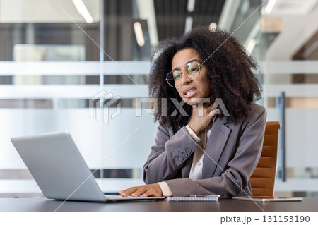 African American young businesswoman sitting in the office at her desk, holding her neck, feeling a strong pain in her throat. 131153190