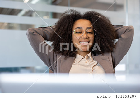 Close-up photo of a young African-American woman in a business suit sitting at a desk in the office, with her hands behind her head and resting contentedly. Close-up photo of a young African-American woman in a business suit sitting at a desk in the office, with her hands behind her head and resting contentedly. 131153207
